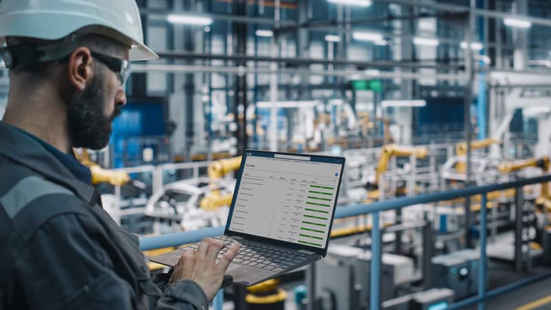 Industriearbeiter mit Schutzhelm nutzt einen Laptop auf einer Produktionshalle-Brücke; im Hintergrund automatisierte Fertigungsanlagen und Roboterlinien.