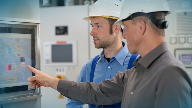 Zwei Industriearbeiter mit Schutzhelmen stehen vor einem Bedienpanel in einer Produktionsumgebung; einer zeigt auf einen Touchscreen mit Prozessdaten und Steuerungselementen.
